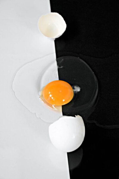 Открытое яйцо с нетронутым желтком в окружении яичных белков лежит на полу-черном полубелом фоне - концепция с черно-белым и яйцом
 