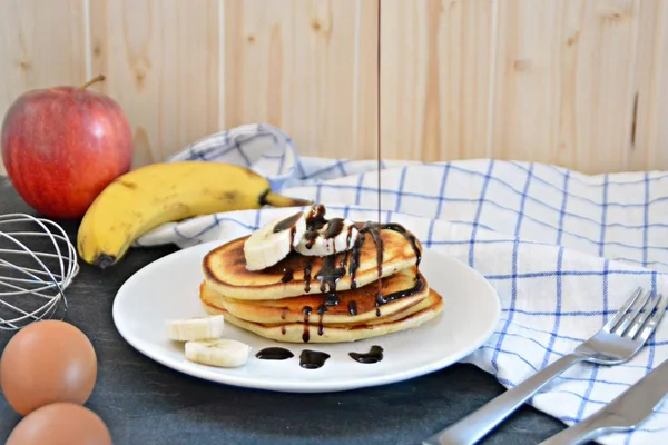 Pancakes with bananas and chocolate sauce on a black table with a wooden background, together with fruit, eggs and a blue white checked kitchen towel