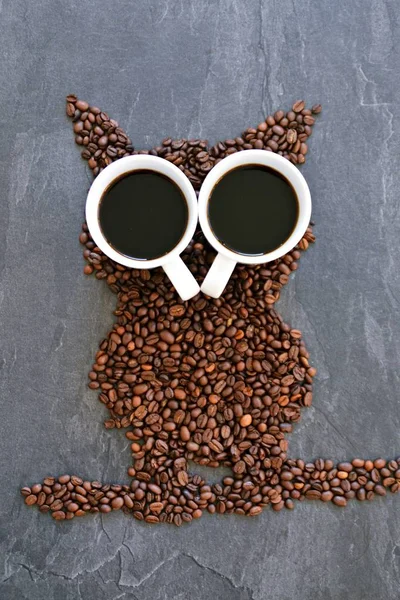 Coffee beans on a black background form the silhouette of an owl and two cups of coffee form their eyes - concept with coffee and night owls