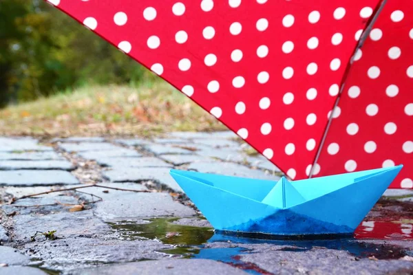 A self-folded paper boat floats in a puddle in nature and is greeted by a pink umbrella with white dots - autumn scenario with paper boat in nature