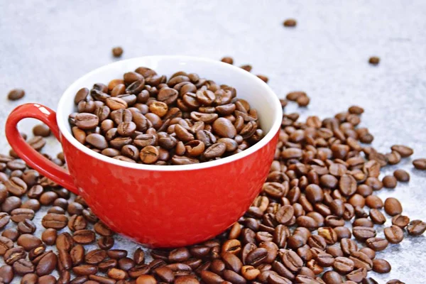 一个装满咖啡豆的红色杯子站在灰色的草坪上 旁边还有其它咖啡豆 这是新鲜咖啡豆品质和享受的概念 — 图库照片