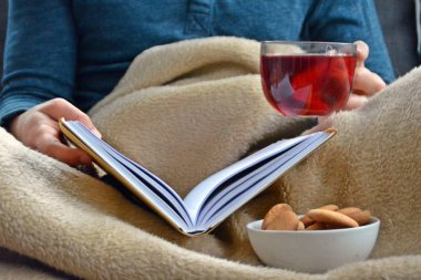 Genç bir adam bir kanepe üzerinde oturuyor ve el ve bisküvi onun yanında stand bir cam bardak bir meyve çayı tutarken bir kitap okuma-kitap ve cam fincan çay dolu-kavramı ısınmak için