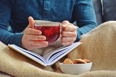 Genç bir adam bir kanepe üzerinde oturuyor ve el ve bisküvi onun yanında stand bir cam bardak bir meyve çayı tutarken bir kitap okuma-kitap ve cam fincan çay dolu-kavramı ısınmak için