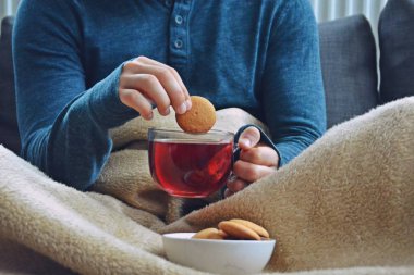 Genç bir adam bir kanepe üzerinde oturuyor ve el ve bisküvi onun yanında stand bir cam bardak bir meyve çayı tutarken bir kitap okuma-kitap ve cam fincan çay dolu-kavramı ısınmak için