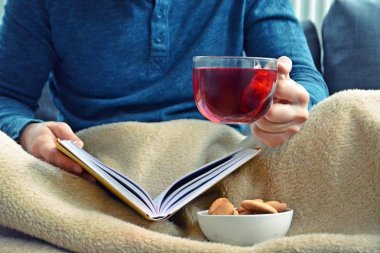 Genç bir adam bir kanepe üzerinde oturuyor ve el ve bisküvi onun yanında stand bir cam bardak bir meyve çayı tutarken bir kitap okuma-kitap ve cam fincan çay dolu-kavramı ısınmak için