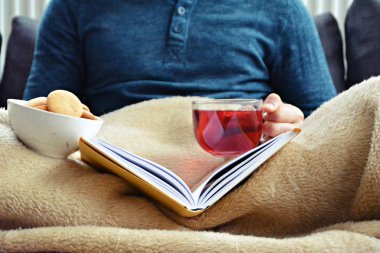 Genç bir adam bir kanepe üzerinde oturuyor ve el ve bisküvi onun yanında stand bir cam bardak bir meyve çayı tutarken bir kitap okuma-kitap ve cam fincan çay dolu-kavramı ısınmak için