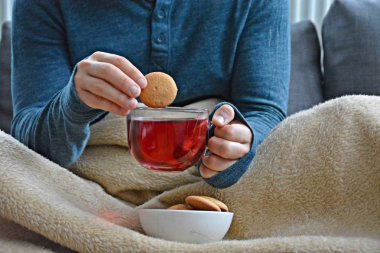 Genç bir adam bir kanepe üzerinde oturuyor ve el ve bisküvi onun yanında stand bir cam bardak bir meyve çayı tutarken bir kitap okuma-kitap ve cam fincan çay dolu-kavramı ısınmak için