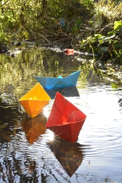 Different colored paper ships swim in a small brook on the surface of which reflects the late autumn sun - Slightly blurred paper ships in bright colors on a lake in late autumn