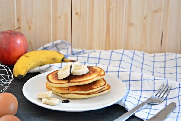 Pancakes with bananas and chocolate sauce on a black table with a wooden background, together with fruit, eggs and a blue white checked kitchen towel