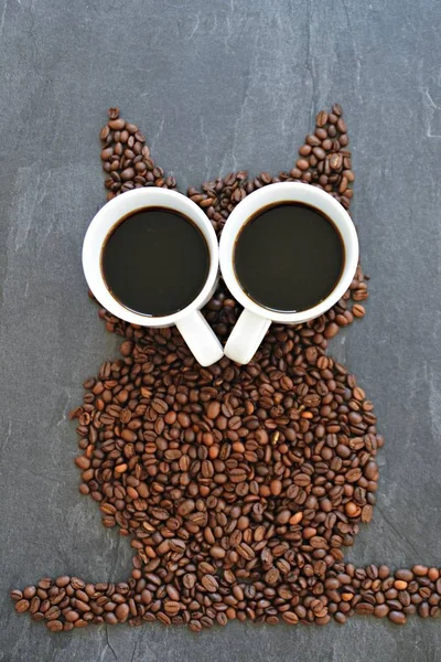 Coffee beans on a black background form the silhouette of an owl and two cups of coffee form their eyes - concept with coffee and night owls