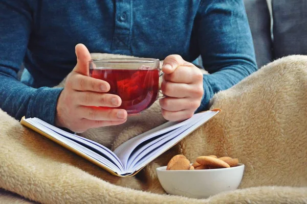 Genç bir adam bir kanepe üzerinde oturuyor ve el ve bisküvi onun yanında stand bir cam bardak bir meyve çayı tutarken bir kitap okuma-kitap ve cam fincan çay dolu-kavramı ısınmak için