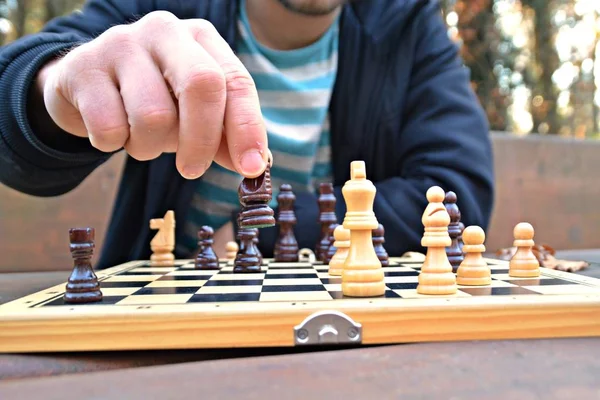 A man in his mid-twenties sits in an autumnal park and plays chess - focusing on the game pieces on the board, the person can not be seen and only the hands and torso are visible