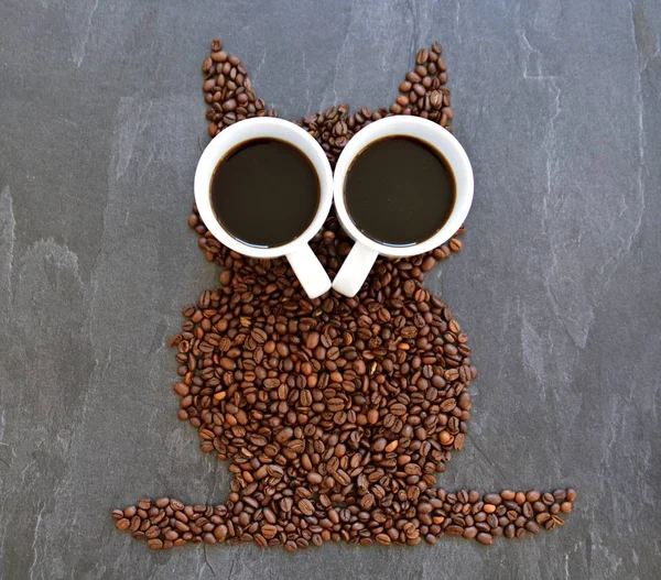 Coffee beans on a black background form the silhouette of an owl and two cups of coffee form their eyes - concept with coffee and night owls