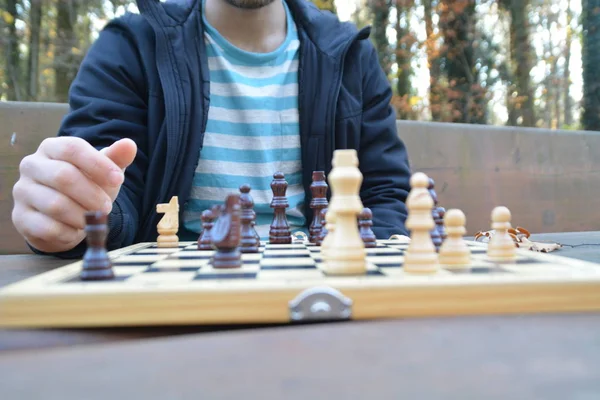 A man in his mid-twenties sits in an autumnal park and plays chess - focusing on the game pieces on the board, the person can not be seen and only the hands and torso are visible