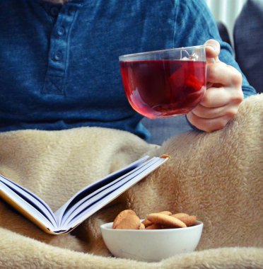 Genç bir adam bir kanepe üzerinde oturuyor ve el ve bisküvi onun yanında stand bir cam bardak bir meyve çayı tutarken bir kitap okuma-kitap ve cam fincan çay dolu-kavramı ısınmak için