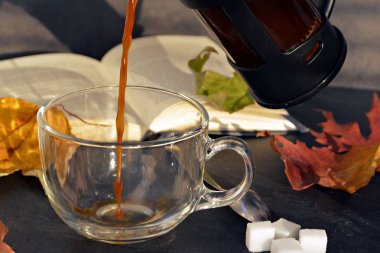 Bir cam fincan içine bir sonbahar ayarı kahve dökün-bir cam fincan kuru ve renkli sonbahar yaprakları ve arka planda bir kitap yanında standig ve birisi içine kahve dökme olduğunu