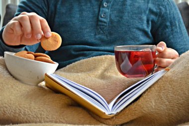 Genç bir adam bir kanepe üzerinde oturuyor ve el ve bisküvi onun yanında stand bir cam bardak bir meyve çayı tutarken bir kitap okuma-kitap ve cam fincan çay dolu-kavramı ısınmak için