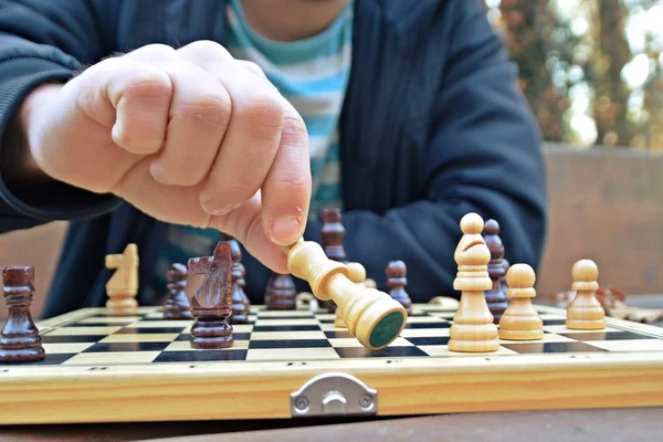 A man in his mid-twenties sits in an autumnal park and plays chess - focusing on the game pieces on the board, the person can not be seen and only the hands and torso are visible