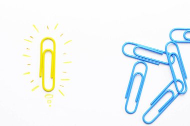 A yellow paper clip as a symbol of a light bulb that radiates lies on a white surface and symbolized the brainstorming - next to it are other paper clips without illusion  clipart