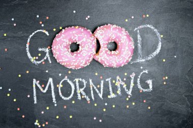 Tebeşir karanlık bir taş yüzeye yazıldı. Günaydın, O kelimeden O kelime beyaz Sprinkles ile iki pembe donut ile değiştirildi-tatlı donut ile güne mutlu başlangıç