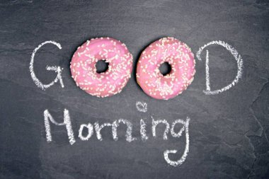 Tebeşir karanlık bir taş yüzeye yazıldı. Günaydın, O kelimeden O kelime beyaz Sprinkles ile iki pembe donut ile değiştirildi-tatlı donut ile güne mutlu başlangıç