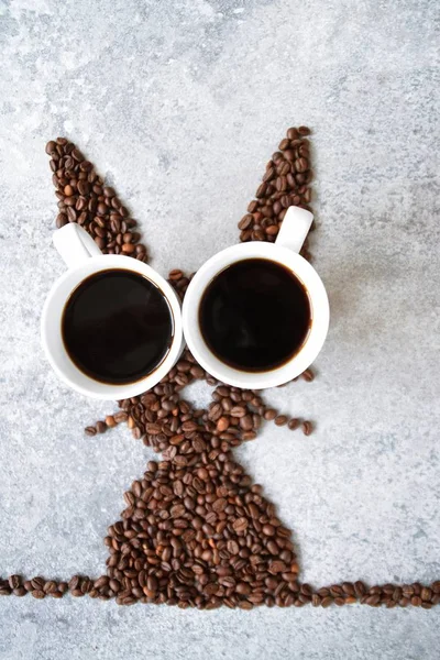 With fresh coffee beans and two cups of freshly brewed black coffee, a hare is shaped on a bright marble surface - concept with coffee beans as a gift for Easter - with room for text or other elements