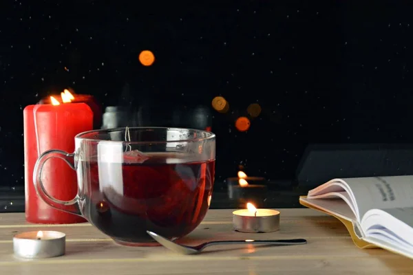 A large glass of fruit tea standing on the wooden windowsill in the night in front of a rain-soaked window in which the lights of the night can be seen - cozy rainy night with a cup of tea