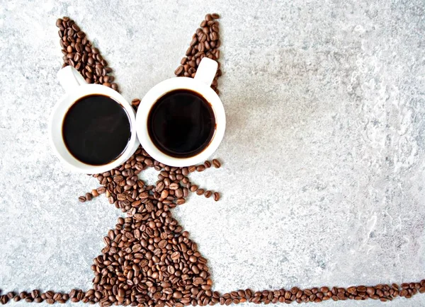 With fresh coffee beans and two cups of freshly brewed black coffee, a hare is shaped on a bright marble surface - concept with coffee beans as a gift for Easter - with room for text or other elements