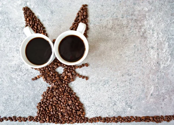 With fresh coffee beans and two cups of freshly brewed black coffee, a hare is shaped on a bright marble surface - concept with coffee beans as a gift for Easter - with room for text or other elements