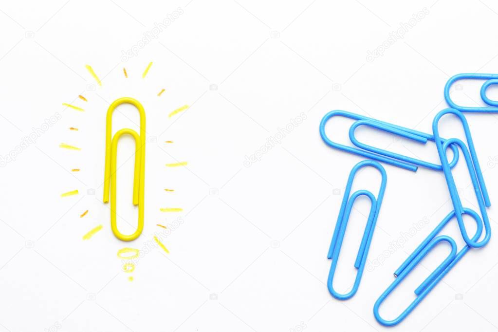 A yellow paper clip as a symbol of a light bulb that radiates lies on a white surface and symbolized the brainstorming - next to it are other paper clips without illusion 