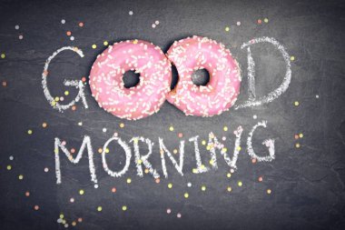 Tebeşir karanlık bir taş yüzeye yazıldı. Günaydın, O kelimeden O kelime beyaz Sprinkles ile iki pembe donut ile değiştirildi-tatlı donut ile güne mutlu başlangıç