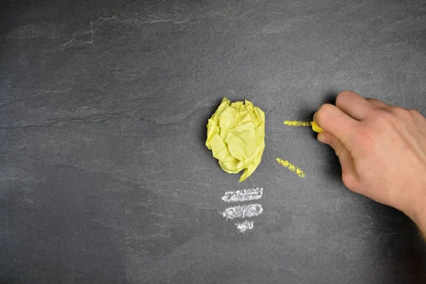A crumpled yellow leaf lies on a dark stone surface and outside was drawn with chalk, a light bulb socket and rays of light - concept for generating ideas and creativity