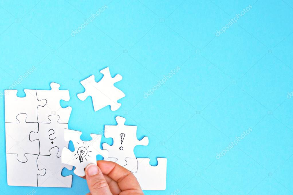 There are puzzle pieces on one face, one on a question mark, the other on an exclamation mark. A hand connects these two parts with a light bulb puzzle piece. Concept for generating ideas with space 