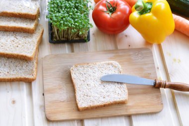 Kaba-taneli krem peyniri ve Cress ile bir kepekli tost bir bıçak yanında bir tahta tahta ve biber ve domates gibi sebze, yanı sıra Cress, salatalık ve havuç bir masada yatıyor