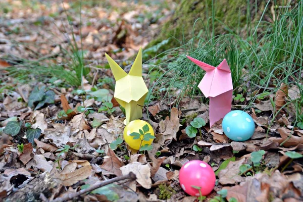 Bunnies folded from colorful paper in origami style stand with a few colorful plastic eggs with white spots on a forest floor in spring