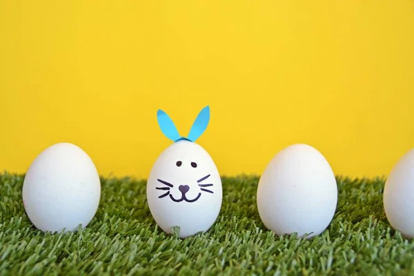 eggs painted with the face of a hare and glued to rabbit ears lie on a grass field in front of a wooden background with space for text or other elements