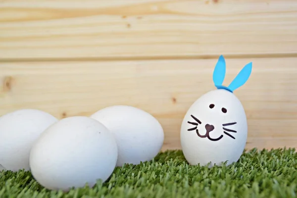 ウサギの顔で塗られ ウサギの耳に接着された卵は テキストや他の要素のためのスペースを持つ木製の背景の前の草原に横たわっています — ストック写真