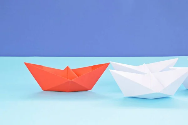 A red self-folded paper boat lies on a blue surface, followed by many white boats - concept symbolizing leadership