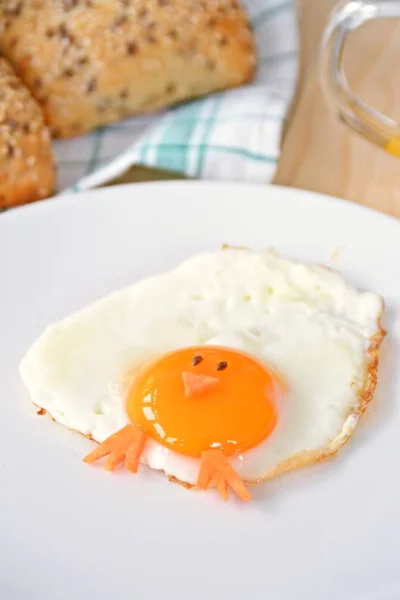 A fried egg lies on a plate, next to it are bread rolls and cutlery. The egg yolk of the fried ice cream looks like a chick, with two grains of eyes and pieces of carrot as mouth and feet