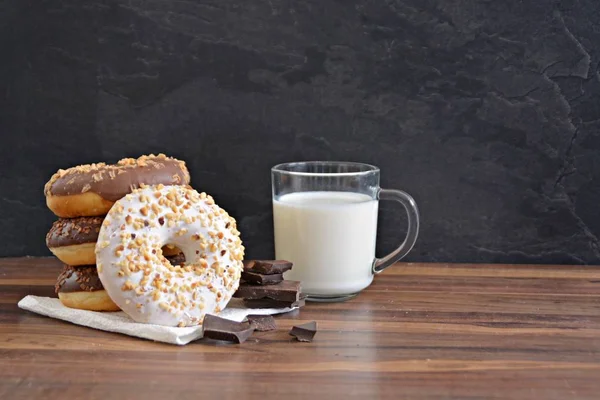 glazed donut with brittle splinters and other chocolate donuts lie on a dark wooden surface against a dark background and a glass of milk - concept for sweet donuts