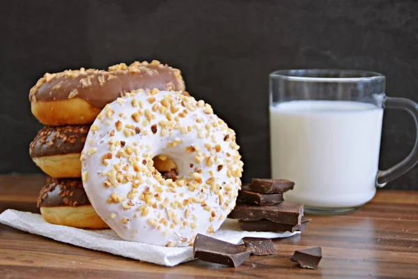 glazed donut with brittle splinters and other chocolate donuts lie on a dark wooden surface against a dark background and a glass of milk - concept for sweet donuts