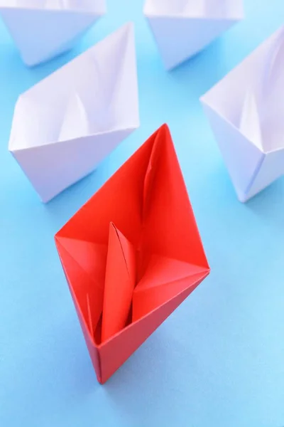 A red self-folded paper boat lies on a blue surface, followed by many white boats - concept symbolizing leadership