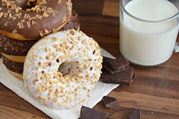 glazed donut with brittle splinters and other chocolate donuts lie on a dark wooden surface against a dark background and a glass of milk - concept for sweet donuts