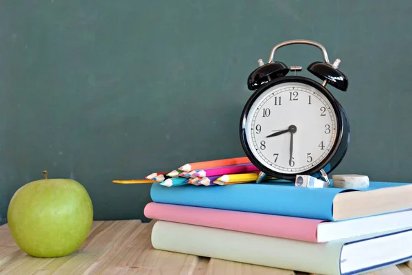 An alarm clock stands in front of a blackboard with colorful books and a green apple - concept with space for text or other elements on the subject of school and time management in elementary school