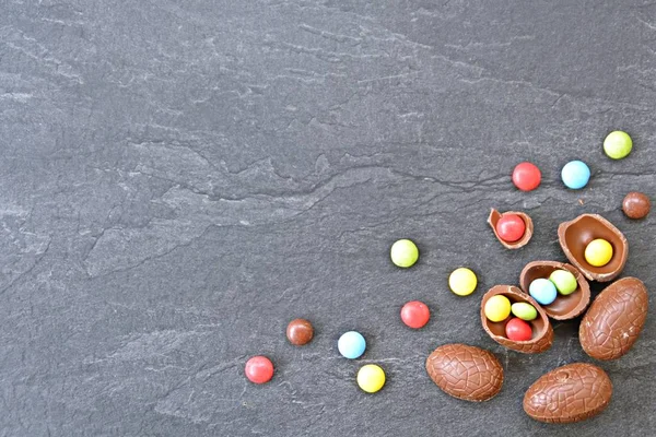 A few chocolate eggs on a dark marble surface, some of which are broken, in the eggs and next to it are colorful chocolate - concept with Easter sweets and space for text or other elements