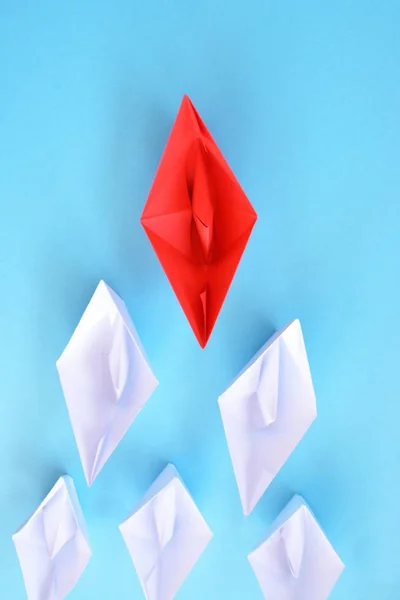 A red self-folded paper boat lies on a blue surface, followed by many white boats - concept symbolizing leadership