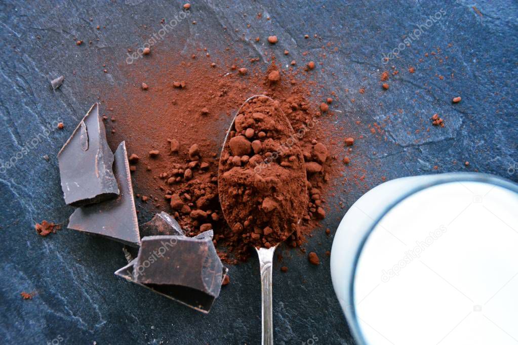 On a dark marble suface lies a spoon full of cacao powder with dark chocolate in fragments next to it 