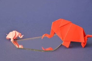 Küçük ve büyük bir origami fil iki fil gibi görünen bir ayna parçası önünde tek renkli bir yüzey üzerinde kırmızı ve pembe kağıt standı katlanmış bir su deliğinin önünde duruyor 