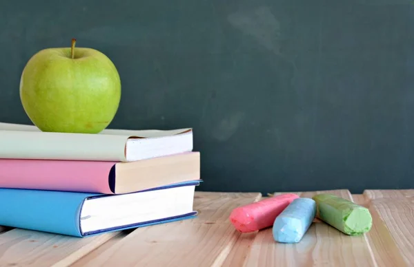 Three books with different colored covers lie on a wooden surface in front of a blackboard with an apple - concept for education and school with place for text or other elements