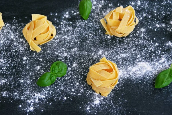 Pasta noodles of the sort tagliatelle lie on a dark marble surface which is lightly sprinkled with flour, next to it are basil leaves - concept as background for fresh prepared noodles